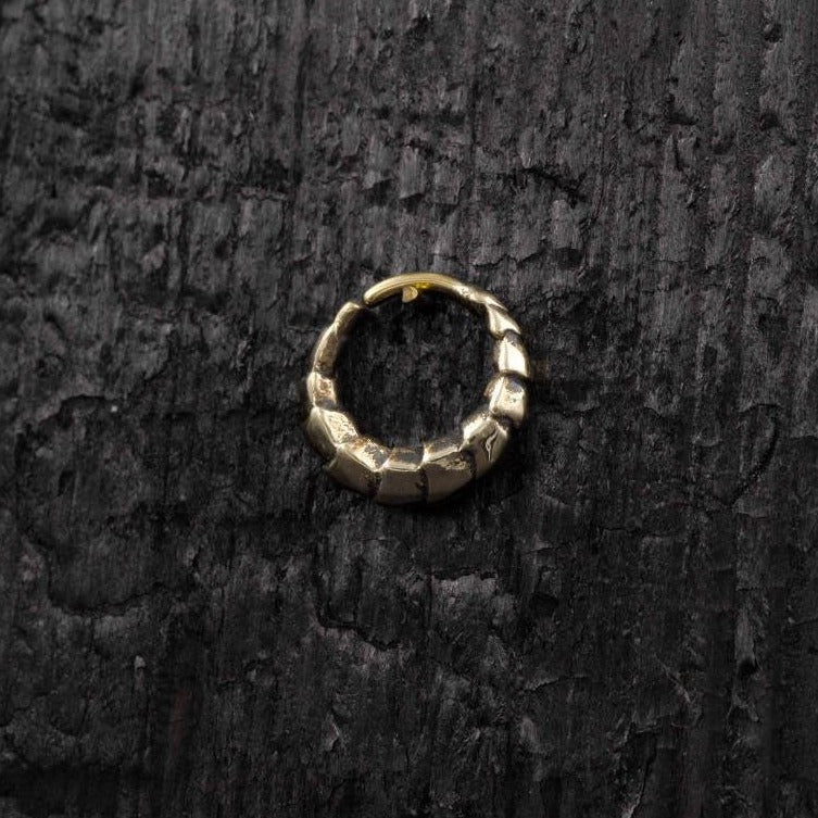 An AARO Cyborg Seamless Hoop Septum Nose Ring in Gold by Kaya Movement, featuring a chunky and rugged design, rests on a dark, rough wooden surface. The 18 gauge gold-colored ring has a closed-loop structure with a clasp-like detail, creating a striking contrast against the dark background.
